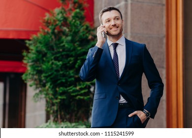 Optimistic Male Owner Of Buisness Company Dressed In Elegant Suit, Has Telephone Conversation, Consults With Operator, Looks Into Distance, Has Happy Look, Stands Outside. People And Communication