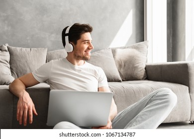 Optimistic Man 30s In Basic Clothing Sitting On Floor At Home And Looking Aside With Smile While Using Laptop And Wireless Headphones