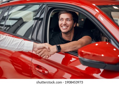 Optimistic man smiling and shaking hand with crop manager while sitting in new red vehicle in dealership