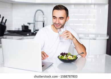 Optimistic man wearing casual clothing eating breakfast and drinking water at home while using laptop