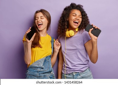 Optimistic pleased mixed race women sing favourite song in smart phones, have fun and enjoy music, keep eyes shut, move actively, isolated on purple background. People, free time and joy concept