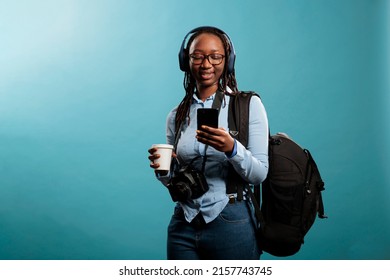 Optimistic Professional Photographer With Smartphone Device, Wireless Headphones And DLSR Camera Listening To Music. Photography Entusiast With Modern Equipment Using Phone To Browse Internet.