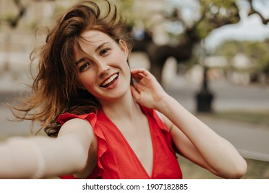 Optimistic Woman In Red Dress Making Selfie Outside. Happy Girl With Sloppy Brown Wavy Hair In Bright Outfit Smiling Outdoors..