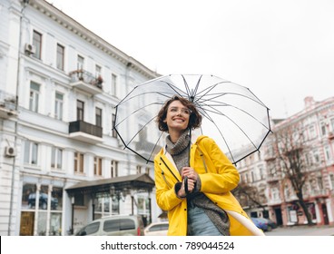 Optimistic woman in yellow raincoat and glasses having fun while walking through city under big transparent umbrella, during cold rainy day - Powered by Shutterstock