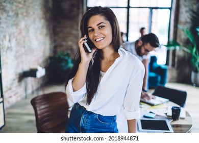 Optimistic Young Woman With Dark Hair Talking On Mobile Phone While Standing In Modern Loft Style Workspace And Looking At Camera