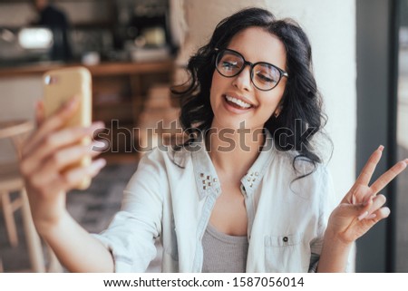 Optimistic young woman gesturing V sign and smiling while taking selfie in cozy restaurant