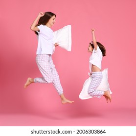 Optimistic young woman and girl with pillows raising arms and leaping up against pink backdrop in morning