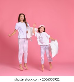 Optimistic young woman and girl with pillows raising arms and leaping up against pink backdrop in morning
