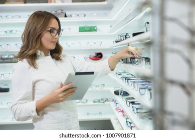 Optometrist is holding tablet computer and looking at glasses at shelf.