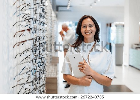 Optometrist is smiling and holding tablet while standing in optical store with many eyeglasses on display and customer choosing eyeglasses in background