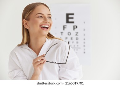 Optometry And Vision. Smiling Girl At Ophthalmologist Office. Portrait Of Beautiful Woman Laughing Out Loud And Holding Glasses With Visual Eye Test Chart On Background. High Resolution Image 