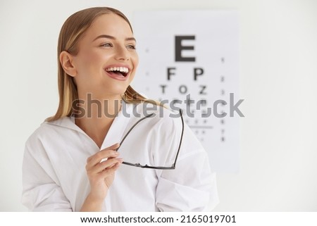 Optometry And Vision. Smiling Girl At Ophthalmologist Office. Portrait Of Beautiful Woman Laughing Out Loud and Holding Glasses with Visual Eye Test Chart On Background. High Resolution Image 