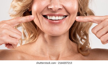 Oral Care. Beautiful Middle Aged Woman Pointing At Her Smile With Two Fingers, Joyful Mature Lady Showing White Teeth, Standing Over Light Grey Studio Background, Cropped Shot, Closeup