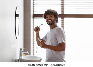 Oral care concept. Happy bearded indian guy in white t-shirt brushing teeth with paste, looking and smiling at camera, standing in modern interior bathroom at home or hotel room