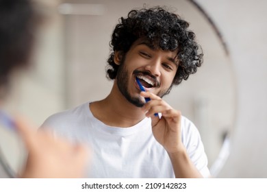 Oral care concept. Young indian man cleaning teeth with toothbrush, smiling to his reflection in mirror, doing toothcare hygiene routine in the morning in bathroom - Shutterstock ID 2109142823
