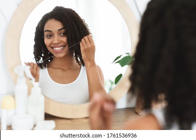 Oral Hygiene. Beautiful african american girl using dental floss at home, looking in the mirror in bathroom, over shoulder view with selective focus