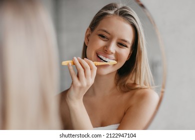 Higiene oral, dientes sanos y cuidado. Mujer joven cepillando dientes con cepillo de dientes y mirando en espejo en el interior del baño por la mañana, armario, espacio vacío
