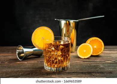 An Orange Alcoholic Cocktail In A Rocks Glass Filled With Ice And Garnished With An Orange Wheel. A Cut Orange, Jigger, Shaker And Strainer. Wooden Table, Dark Grey Background. Studio Horizontal Shot.