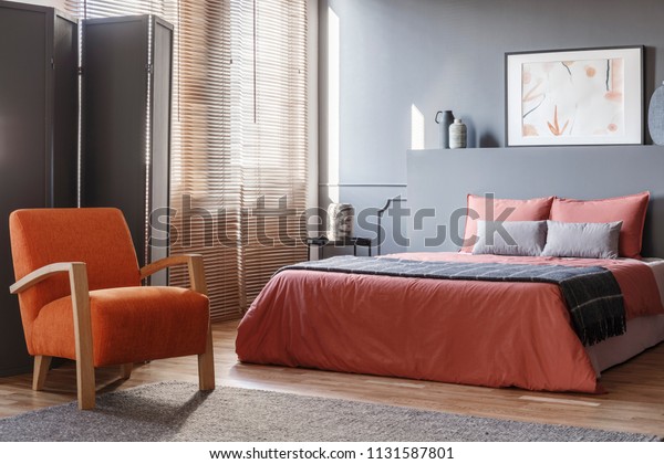 Orange armchair next to pink bed in\
modern grey bedroom interior with screen and\
poster