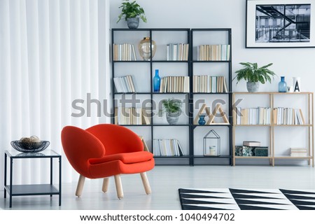 Orange armchair next to table in bright living room interior with bookshelf and poster on white wall