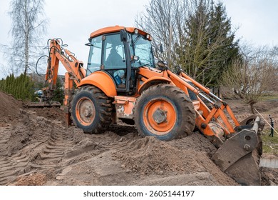 An orange backhoe loader operates on a dirt mound with its rear digging arm extended. Bare trees and sparse vegetation suggest a colder season.