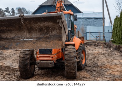 An orange backhoe loader with raised bucket on muddy ground, surrounded by tire tracks. A blue house, metal fence, and trees are visible in the background.