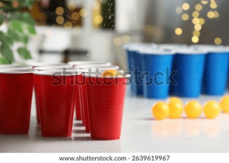 Orange ball falling into plastic cup on light table against blurred lights, closeup. Beer pong game