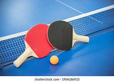 Balón naranja para el ping-pong y dos raquetas de madera de color rojo y negro sobre una mesa azul con rejilla