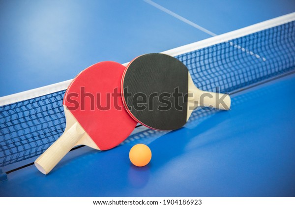 Orange ball for table tennis\
and two wooden rackets of red and black color on a blue table with\
a grid