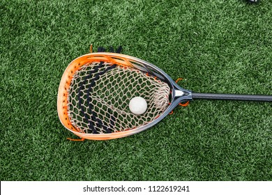 Orange And Black Lacrosse Goalie Stick With A Ball In The Net Lying On A Green Turf Field. 