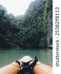 The orange bow of an inflatable canoe, on the smooth water surface of the bay, in front of the bow of the canoe lies an island with steep cliffs and green jungle.