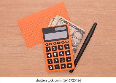 Orange Calculator On US Currency And Index Card Next To Black Felt Tip Pen On Wood Grain Desk