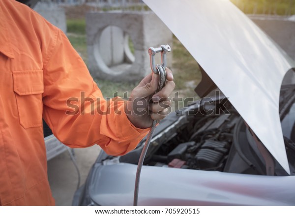 An orange car repairman standing, Holding a\
towing sling pulling a broken\
car.