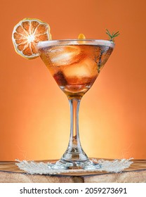 An orange cocktail in martini glass with dehydrated lemon and rosemary sprig on rustic wooden surface