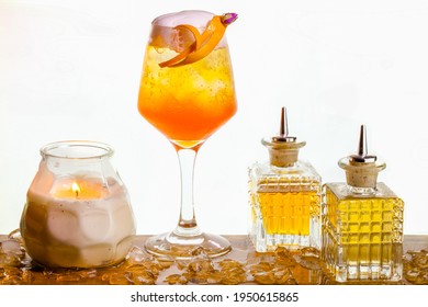 Orange Cocktail Set On A Wooden Surface With A Candle Jar And 2 Small Decorative Bottles, In Front Of A White Background