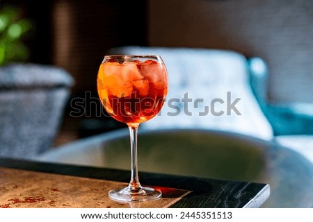 An orange cocktail in a wine glass on a rustic table, with a blurred lounge setting in the background. The vibrant drink features ice cubes and may be an Aperol Spritz.