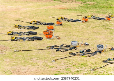 Orange colored half dome helmets arranged in a circle on yellowed lawn with Swift Quick Lock carabiners, handlebars and full body harnesses used for zip lining outdoor sports activities in a camping.