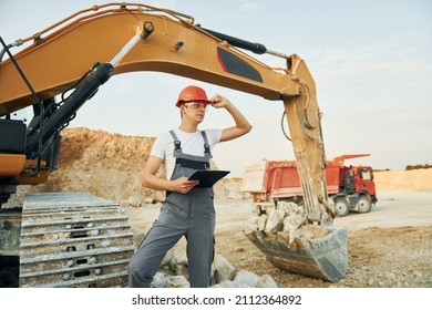 In orange colored hard hat. Worker in professional uniform is on the borrow pit at daytime.