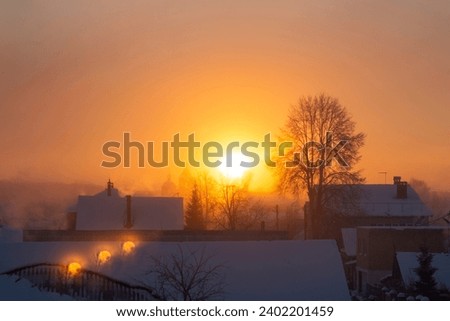 the orange dawn of the sun against a background of trees, roofs of houses and cold fog