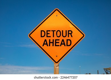 An orange diamond shaped sign stating Detour Ahead against a blue sky