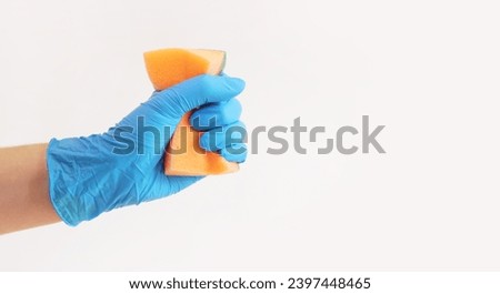 Orange dishwashing sponge in a woman's hand on a gray background. A hand in a latex blue glove holds a sponge for wet cleaning. Professional cleaning. Copy space. The hand squeeze the sponge