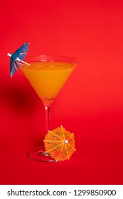 Orange Drink With A Blue Umbrella In A Martini Glass Set Against A Solid Red Background. An Orange Umbrella Lies At The Base Of The Glass.