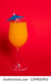 Orange Drink With A Blue Umbrella In A Wine Glass Set Against A Solid Red Background.