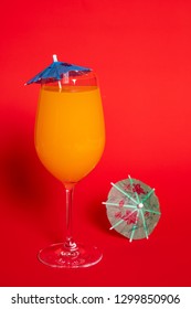 Orange Drink With A Blue Umbrella In A Wine Glass Set Against A Solid Red Background. A Green Umbrella Lies Beside The Glass.