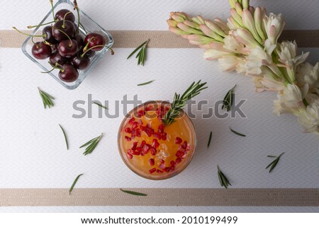 orange drink with red pomegranate and a branch of rosemary, on a table accompanied by cherries and a flower