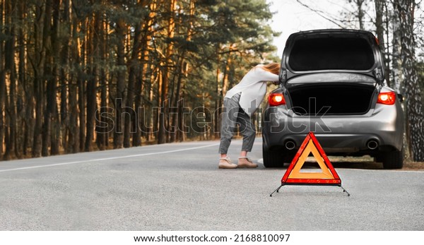 An orange emergency triangle on the road against\
the background of a blurry car and a woman in despair leaning on\
the car waiting for help. Focus on the emergency sign. Broken car\
on the road.