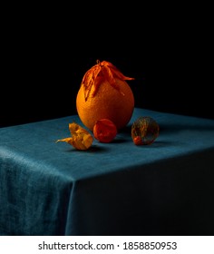 Orange With Flower Crown Stands On Table Next To Dried Flower Buds