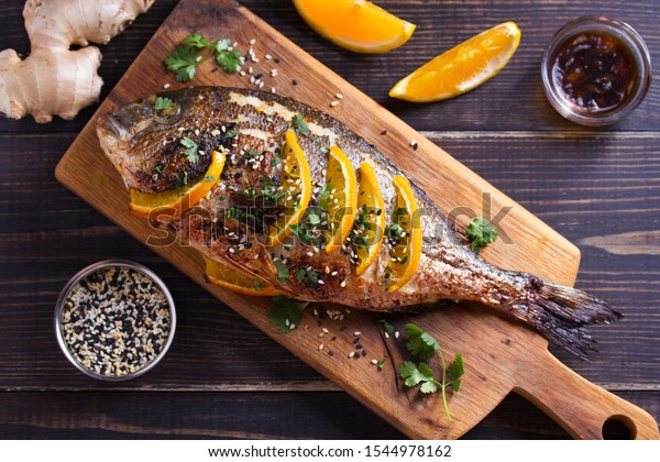 Orange ginger sea bream fish, sprinkled with sesame\
seeds and cilantro on serving board. Soy orange sauce. Overhead\
horizontal image
