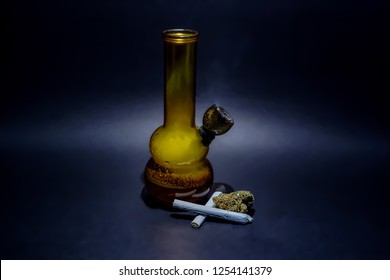 An orange glass water bong full of smoke and dark water with a burning bowl with a small pile of Cheese OG marijuana buds next to it & 2 joints criss crossed isolated with black.