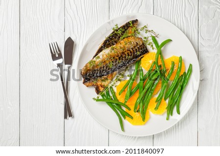 orange glazed mackerel fillet with fresh orange and steamed green beans on a white plate with cutlery on a table, flat lay, free space